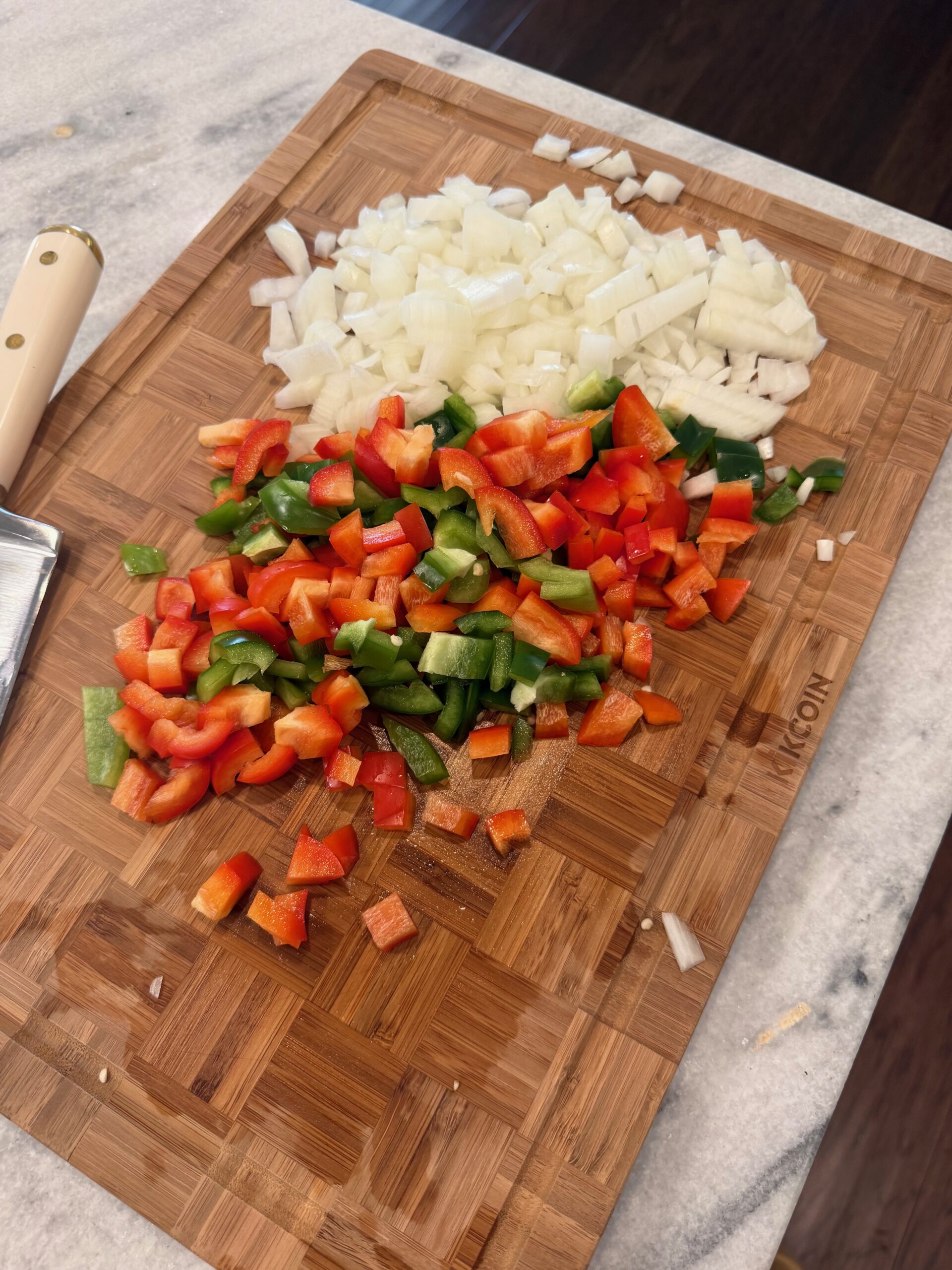 Chopped onions, red bell peppers, and green bell peppers on a wooden cutting board next to a knife on a marble countertop.
