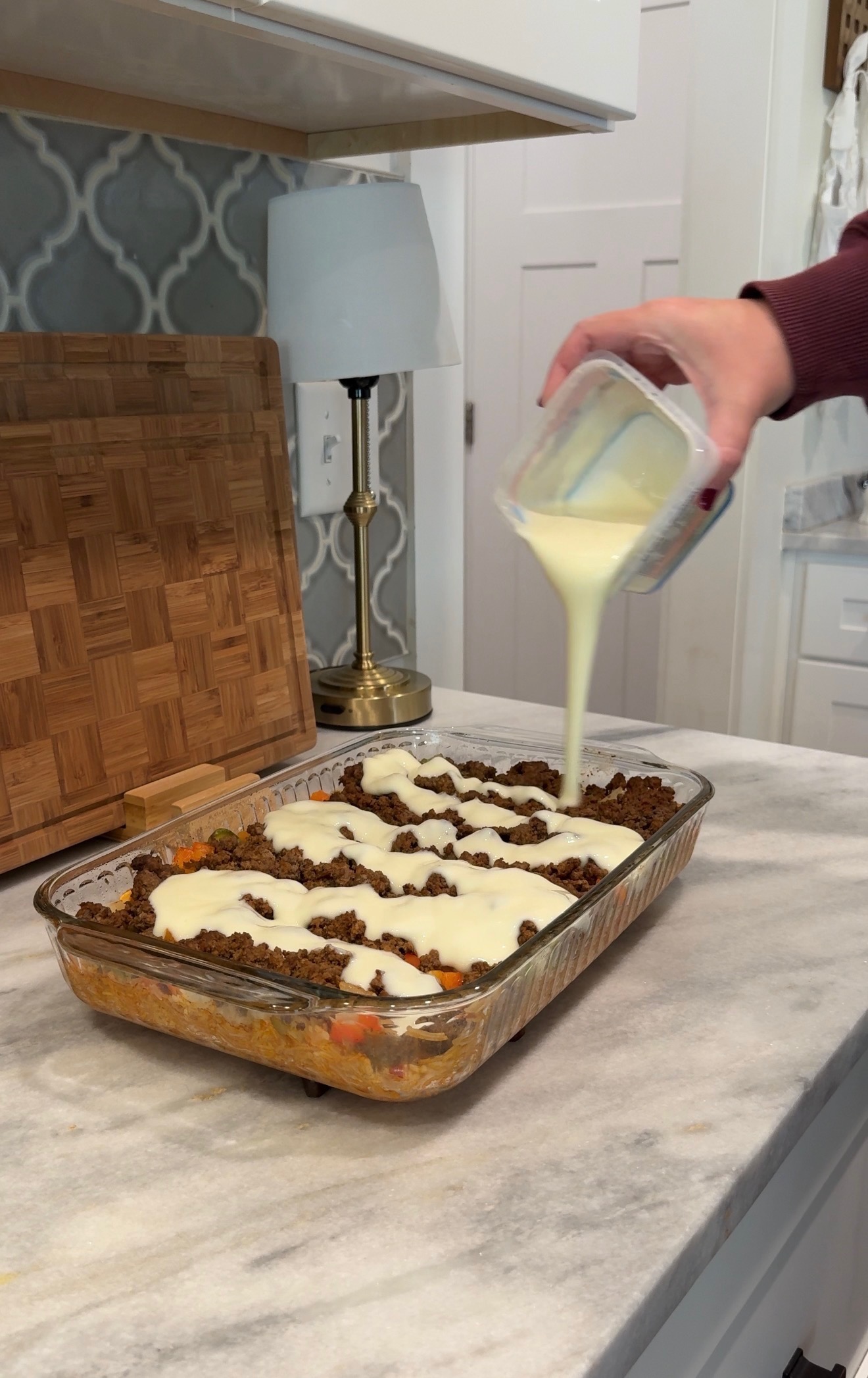 A hand pours creamy white sauce over a casserole topped with ground meat in a glass baking dish on a marble kitchen counter.