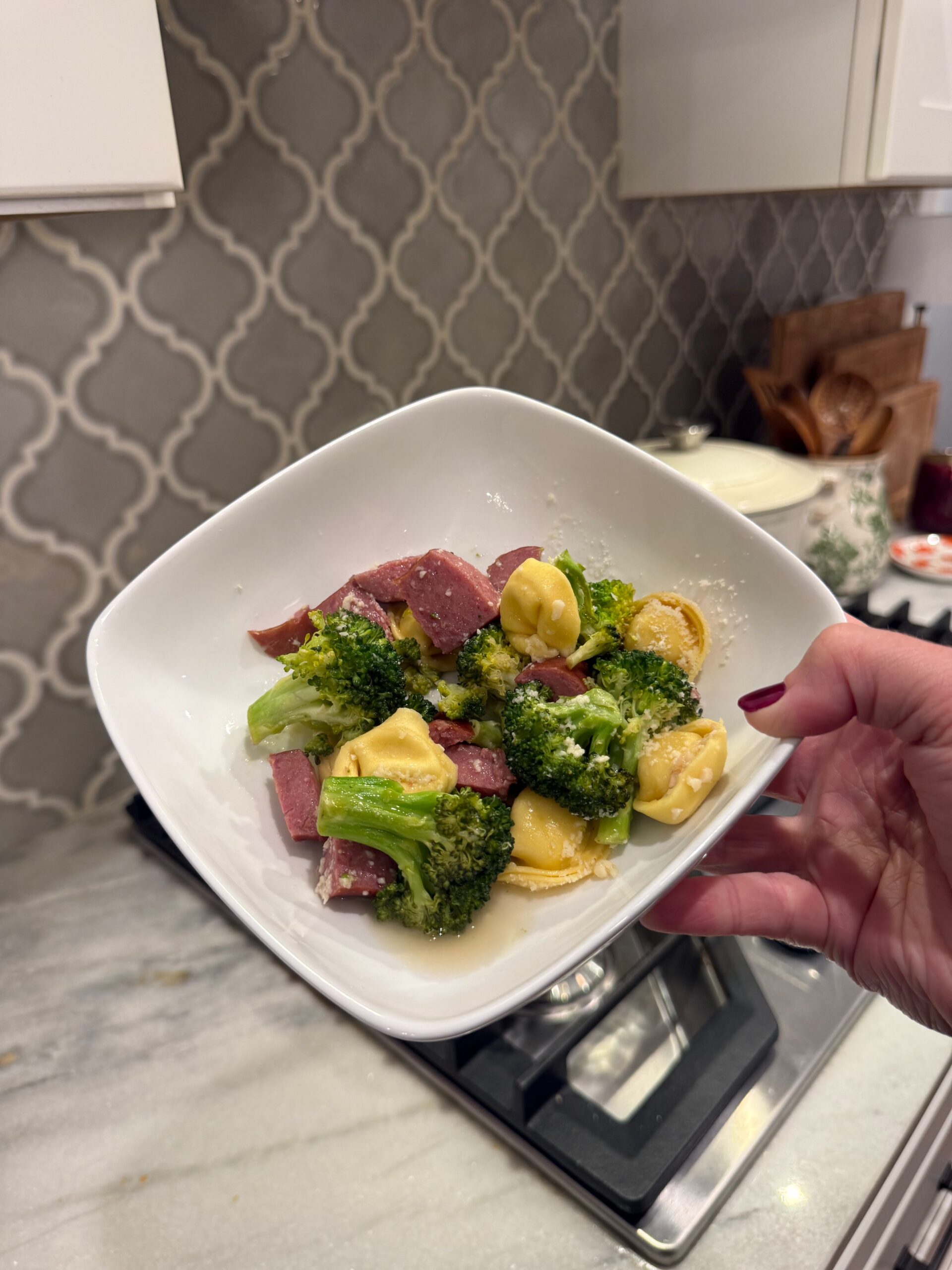 A hand holds a white bowl containing broccoli, tortellini, and slices of meat in a kitchen with a gray tile backsplash and marble countertop.