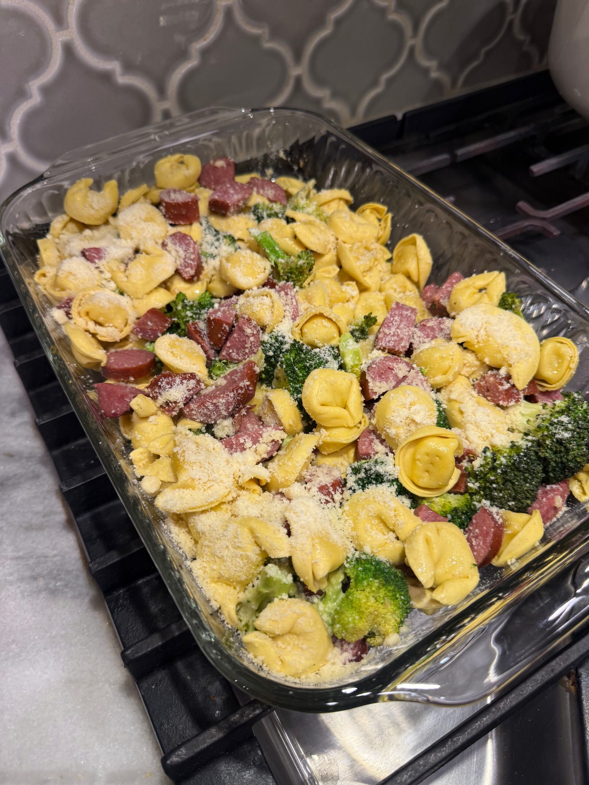 A glass baking dish filled with uncooked tortellini, broccoli, and sausage pieces, sprinkled with grated cheese, sits on a stovetop with a patterned gray tile backsplash in the background.