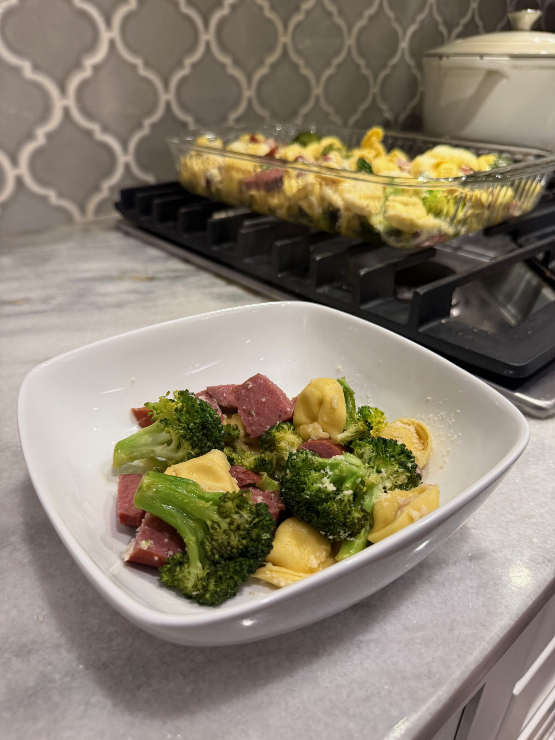 A white bowl filled with tortellini pasta, broccoli, and pieces of sausage sits on a kitchen counter. In the background, a casserole dish with the same meal rests on the stovetop.