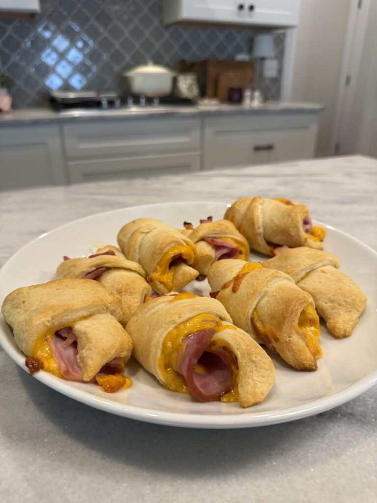 A white plate with baked crescent rolls filled with ham and melted cheese sits on a marble countertop in a kitchen with white cabinets and a tiled backsplash.