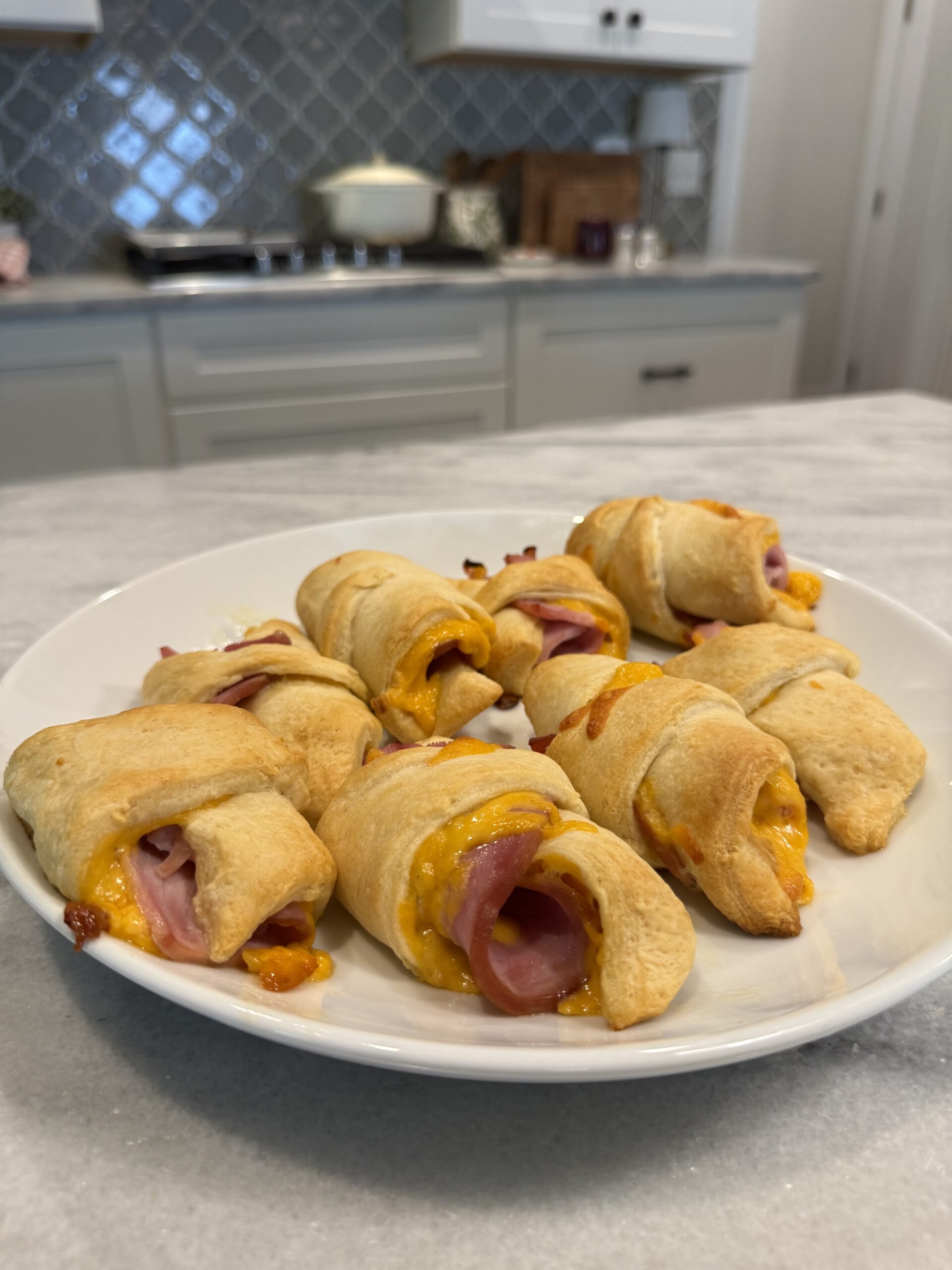 A white plate with baked crescent rolls filled with ham and melted cheese sits on a marble countertop in a kitchen with white cabinets and a tiled backsplash.