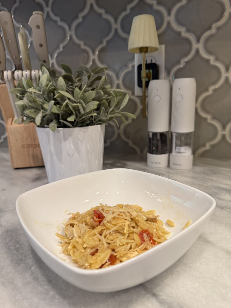 A chicken orzo sits on a marble counter. Behind it are a potted plant, knife block, salt and pepper grinders, and a small wall lamp against a patterned tile backsplash.