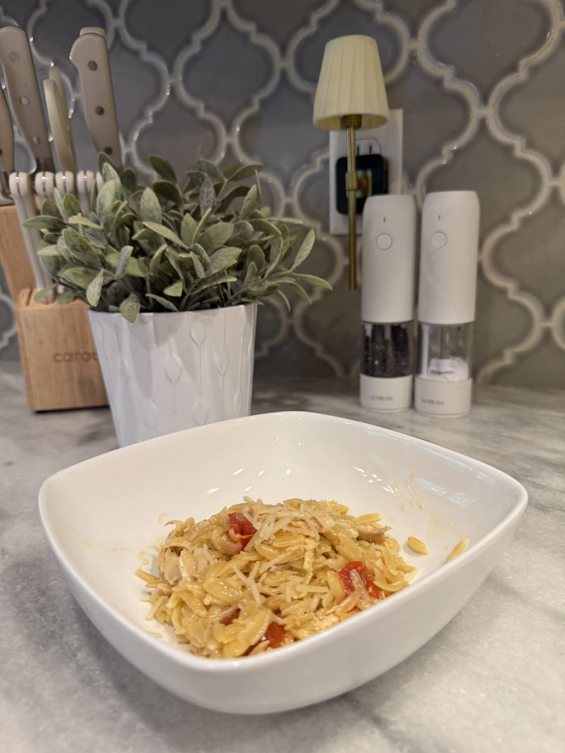 A chicken orzo sits on a marble counter. Behind it are a potted plant, knife block, salt and pepper grinders, and a small wall lamp against a patterned tile backsplash.