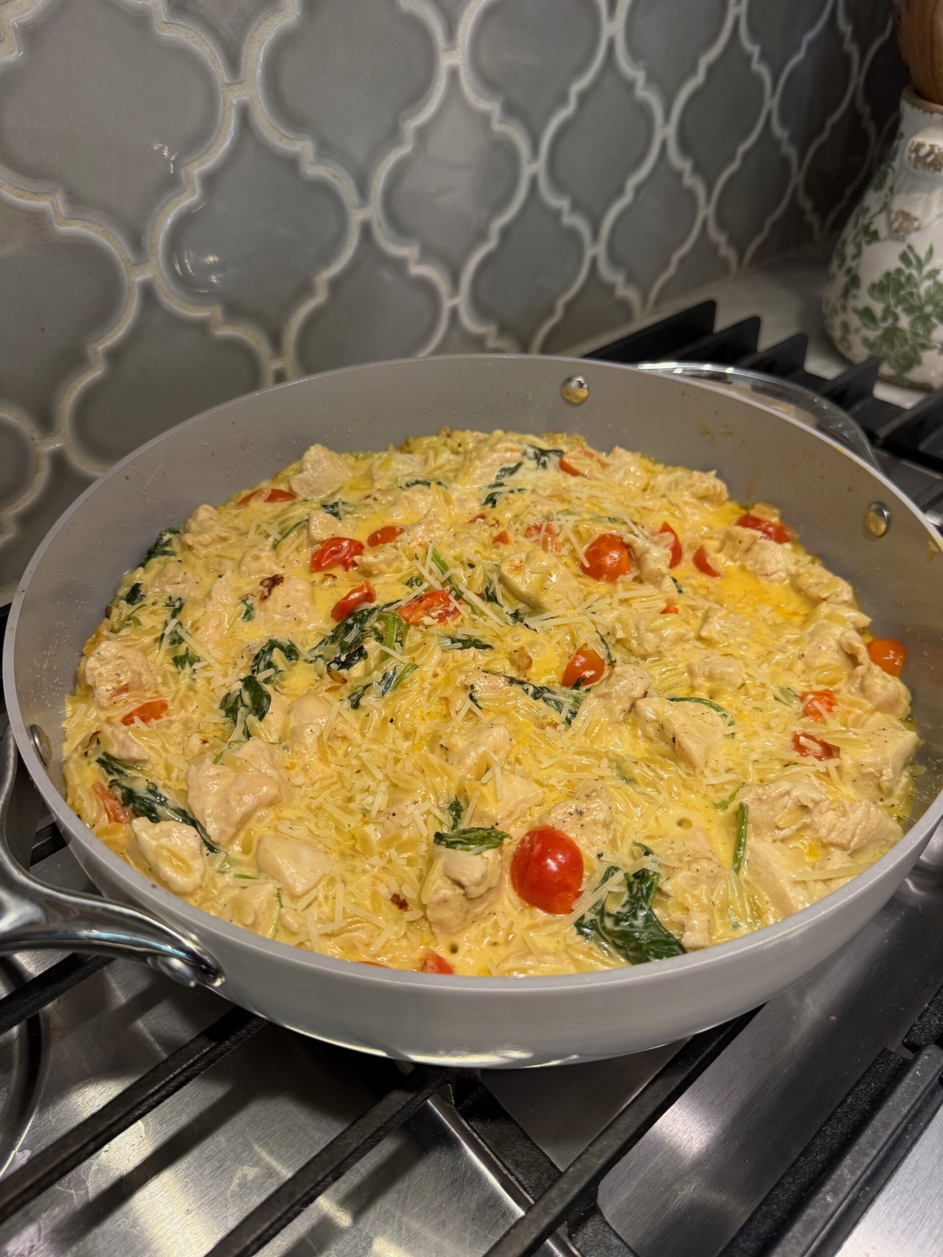 A skillet on a stovetop filled with creamy pasta, chunks of chicken, cherry tomatoes, and spinach. The dish sits against a patterned tile backsplash.