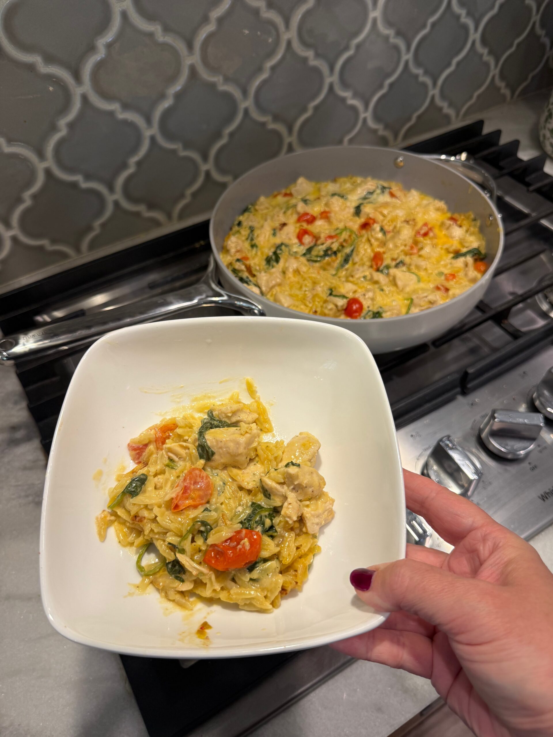 A hand holds a square white bowl with creamy pasta, chicken, spinach, and cherry tomatoes, while a pan with more of the same dish sits on a stove in the background.
