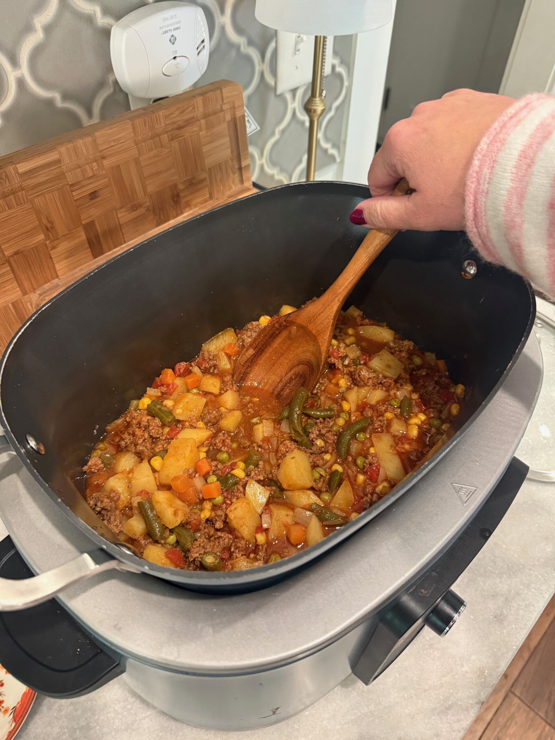 A hand stirs a hearty stew with vegetables and ground meat in a large electric skillet on a kitchen counter.