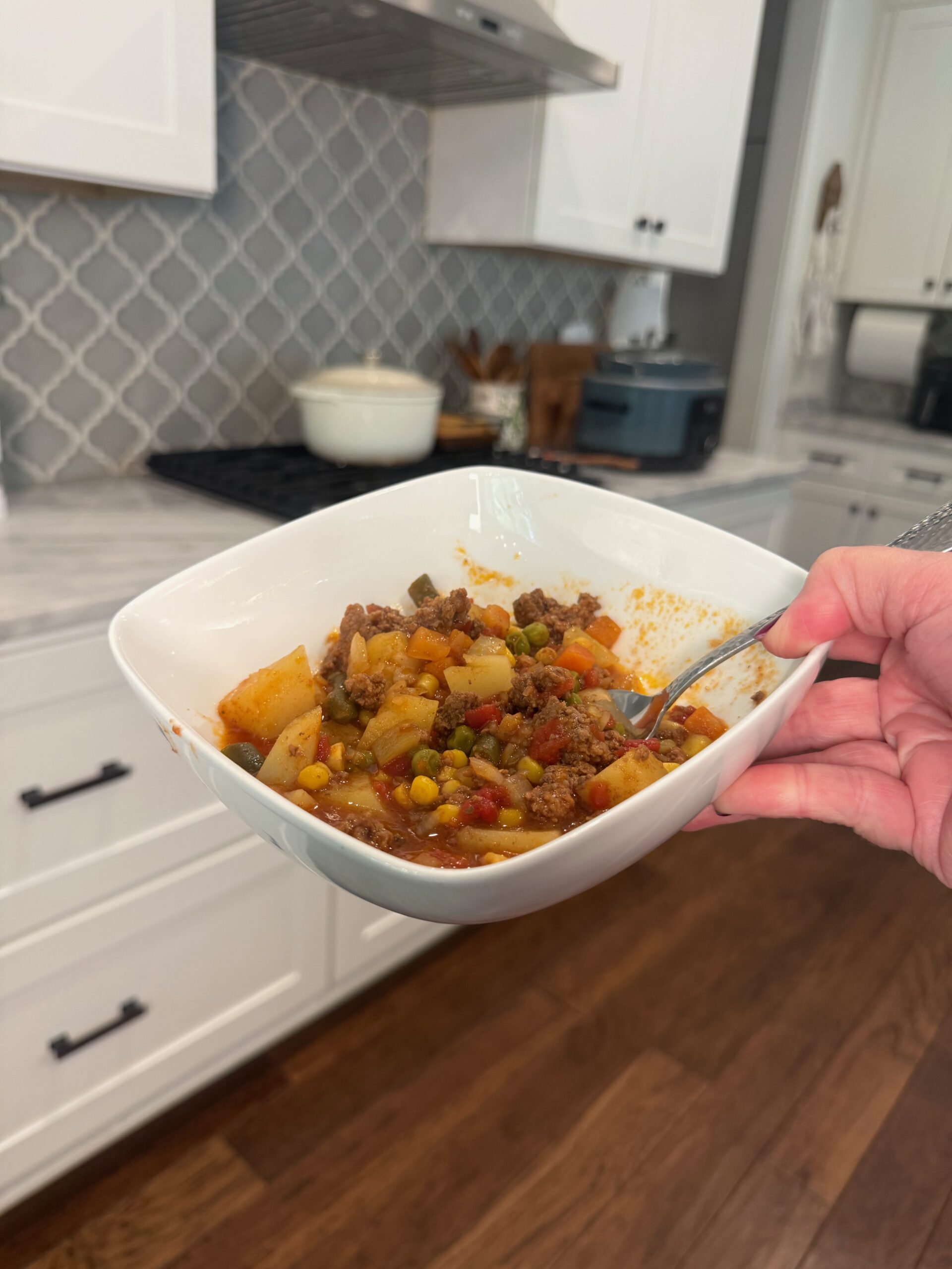 A hand holds a bowl of hearty beef stew with vegetables in a modern kitchen with white cabinets and wood floors.