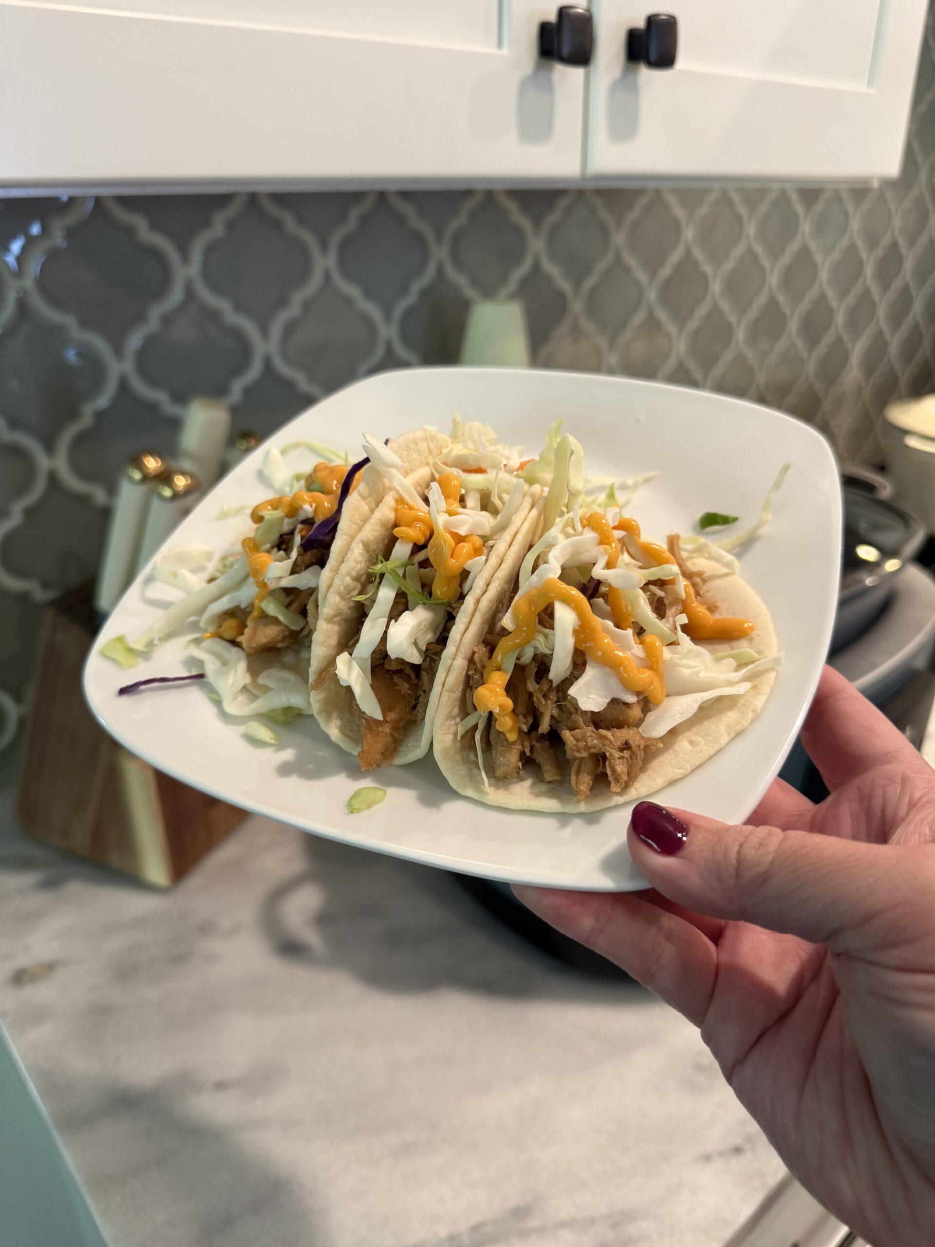 A hand holds a white plate with three tacos topped with shredded cabbage and cheese. The background shows a kitchen counter, backsplash, and cabinets.