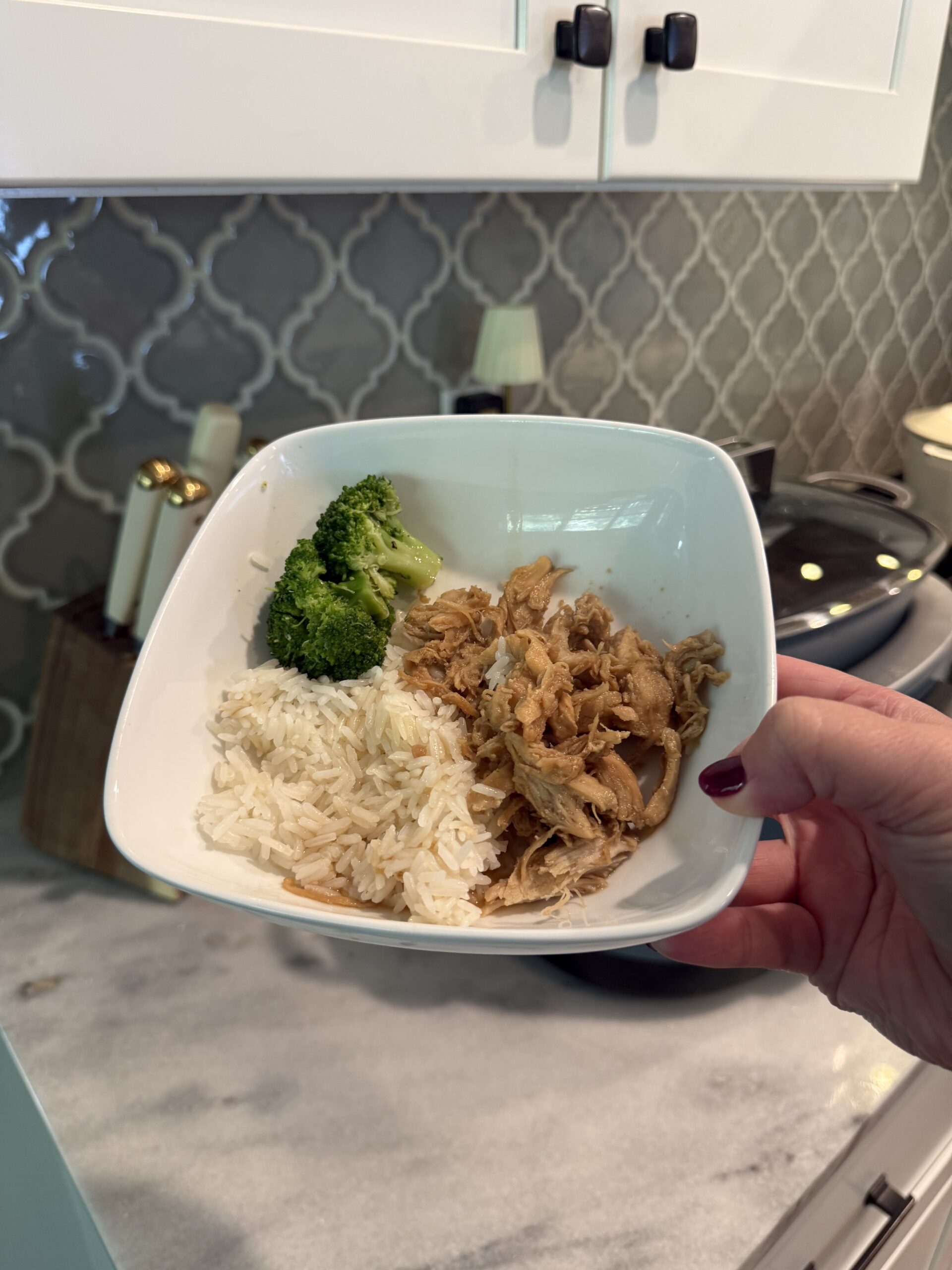 A hand holds a white bowl with shredded chicken, white rice, and steamed broccoli in a kitchen with a gray tile backsplash.