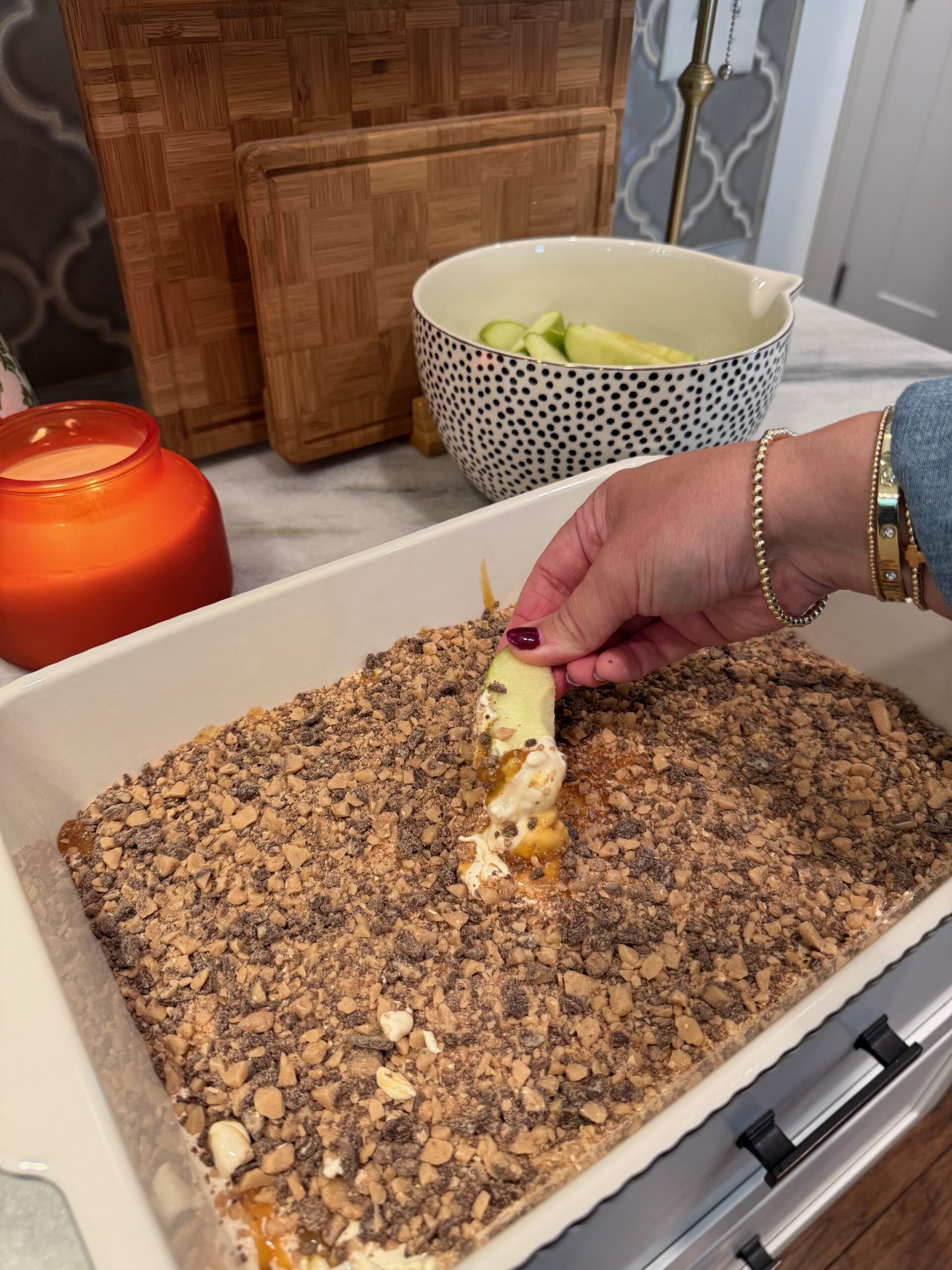 A hand dips a green apple slice into a dessert topped with crushed toffee bits in a white baking dish. A candle, wooden boards, and a bowl of cucumber slices are on the countertop in the background.