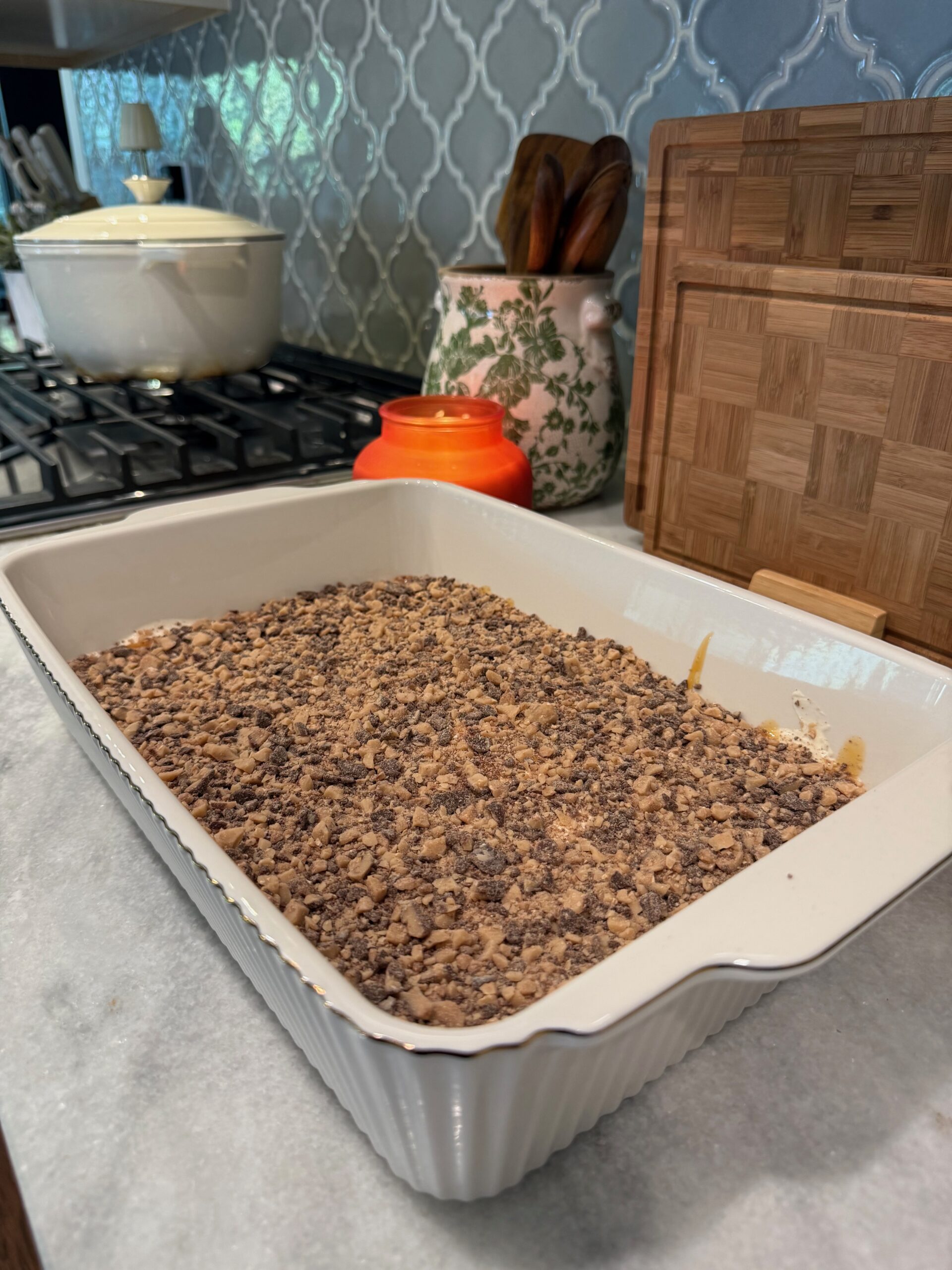 A white baking dish filled with a crumbled topping dessert sits on a kitchen counter. Nearby are a lit orange candle, a green and white utensil holder, and wooden cutting boards.
