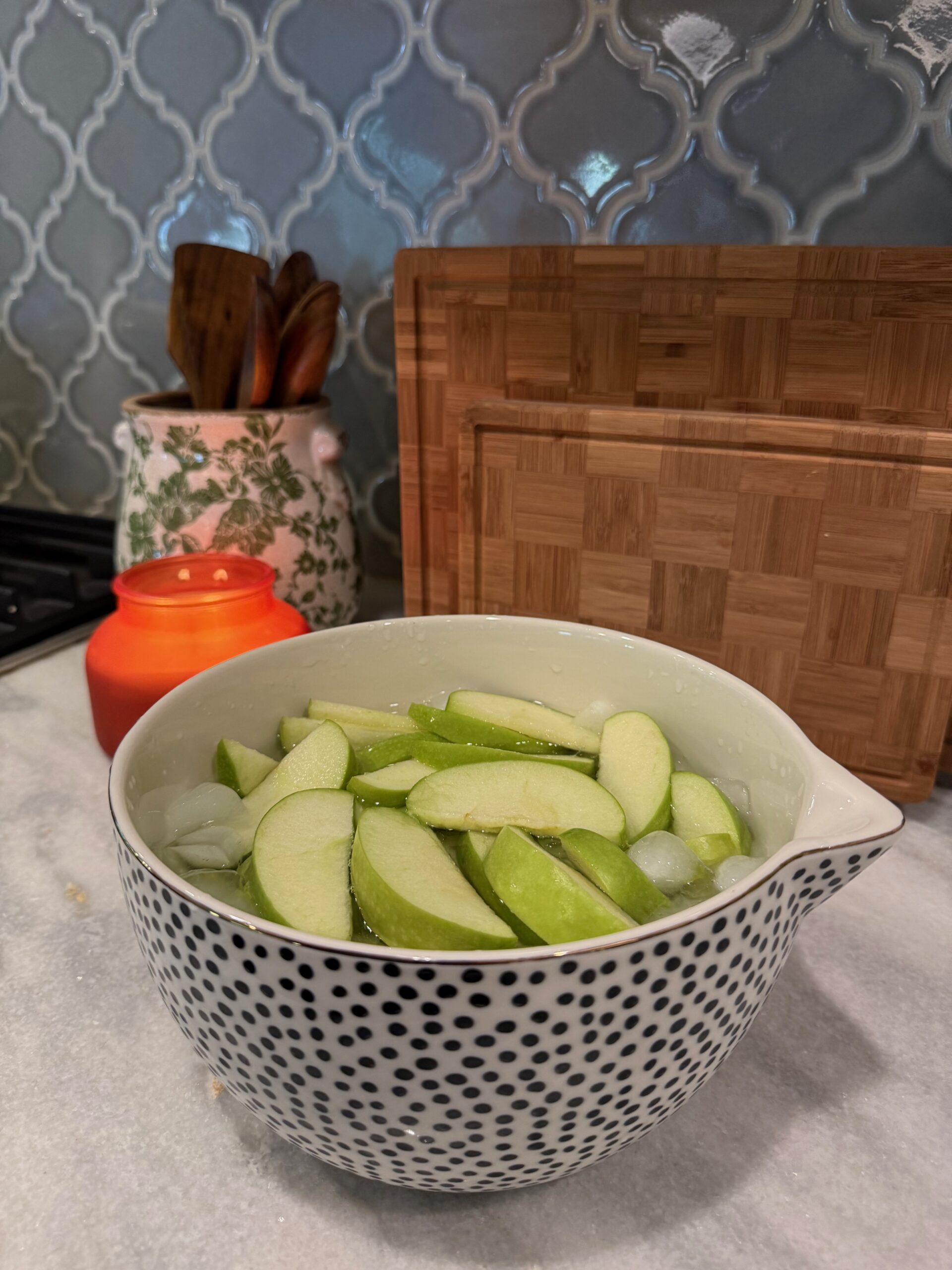 A black and white polka dot bowl filled with sliced green apples sits on a kitchen counter next to a lit orange candle and wooden cutting boards.