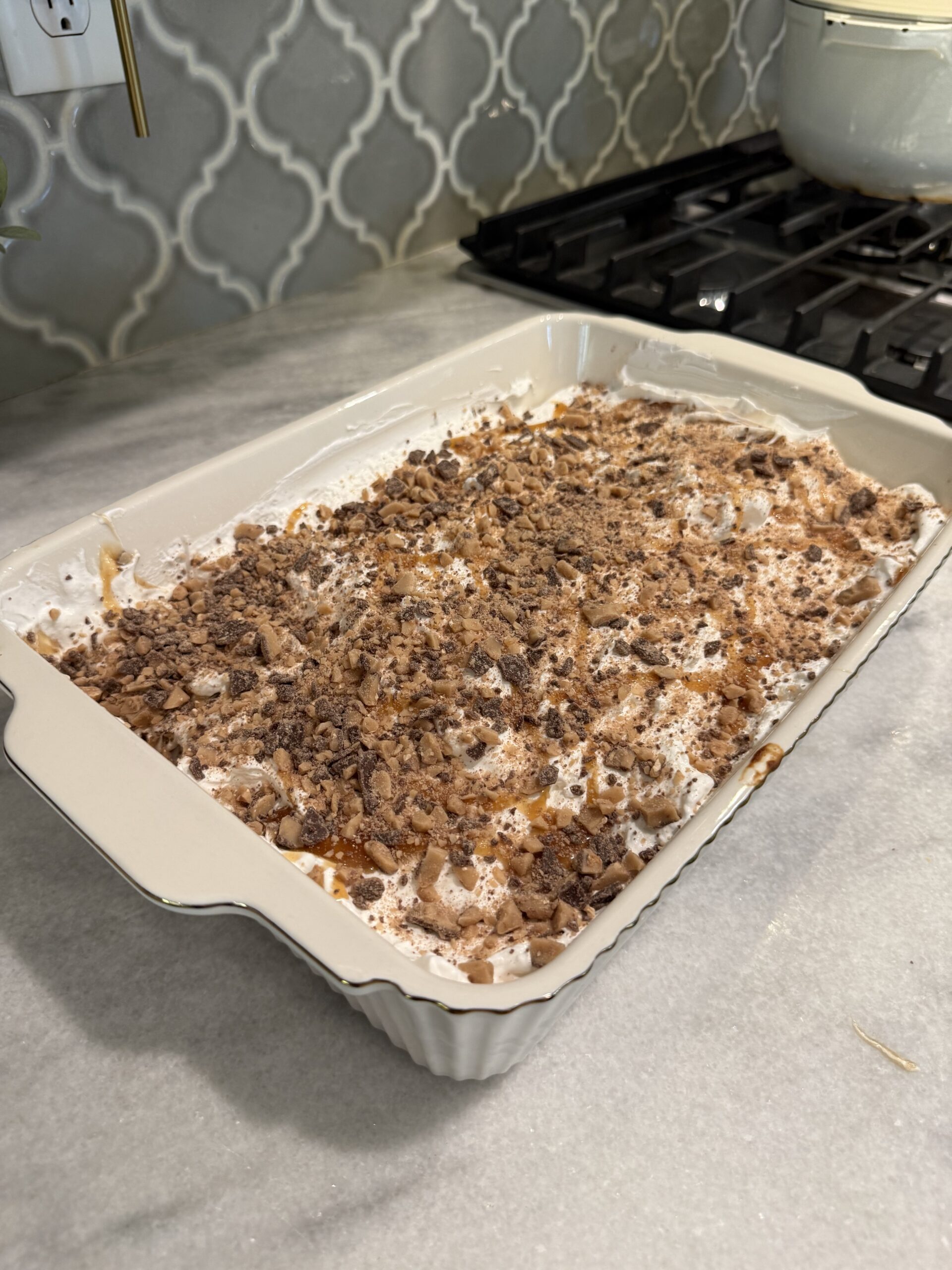 A rectangular baking dish filled with dessert, topped with whipped cream and crushed toffee pieces, sits on a kitchen counter near a stove with a white pot.