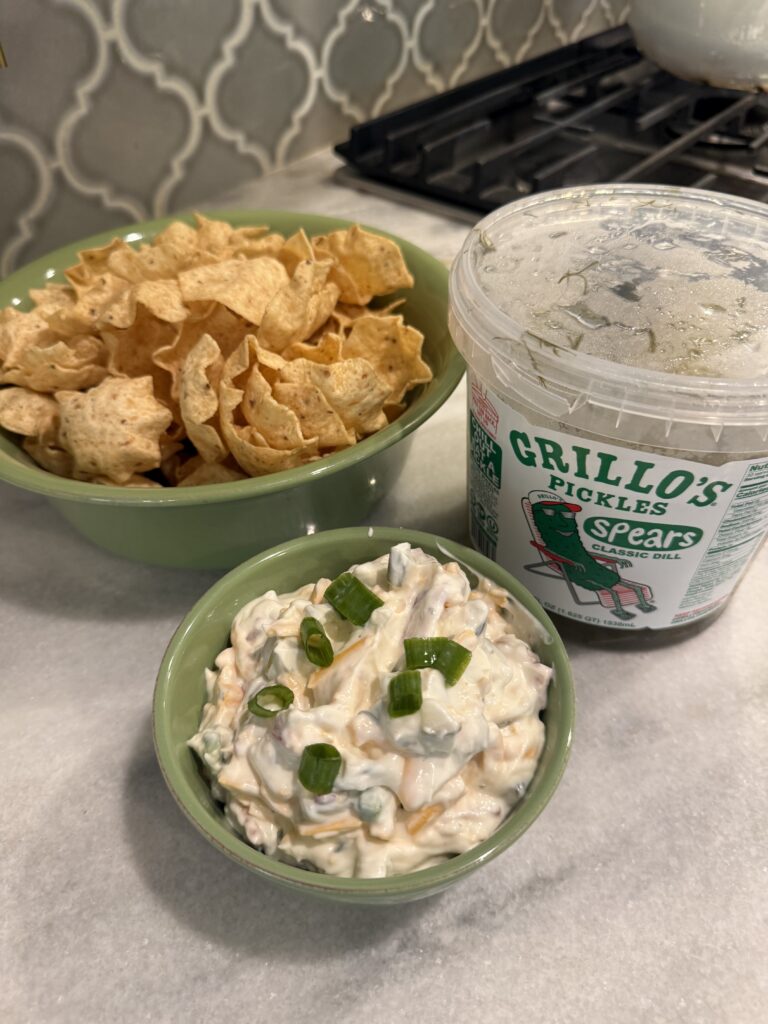A bowl of tortilla chips, a tub of Grillo’s Pickles Spears, and a bowl of creamy pickle dip topped with chopped green onions are arranged on a kitchen counter—an irresistible spread inspired by the viral snack trend.