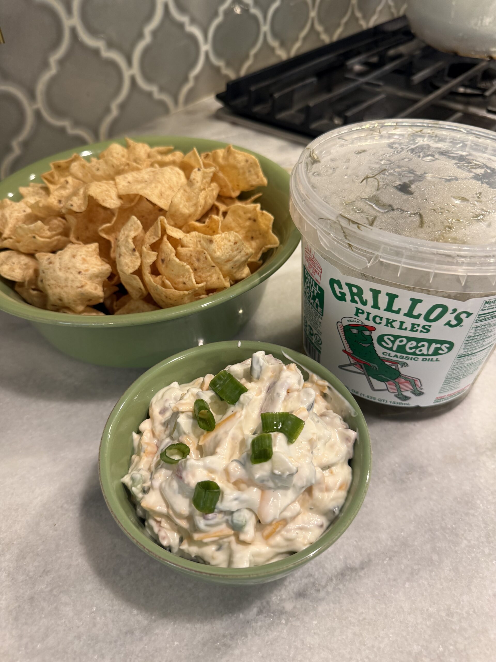 A bowl of tortilla chips, a tub of Grillo’s Pickles Spears, and a bowl of creamy pickle dip topped with chopped green onions are arranged on a kitchen counter—an irresistible spread inspired by the viral snack trend.
