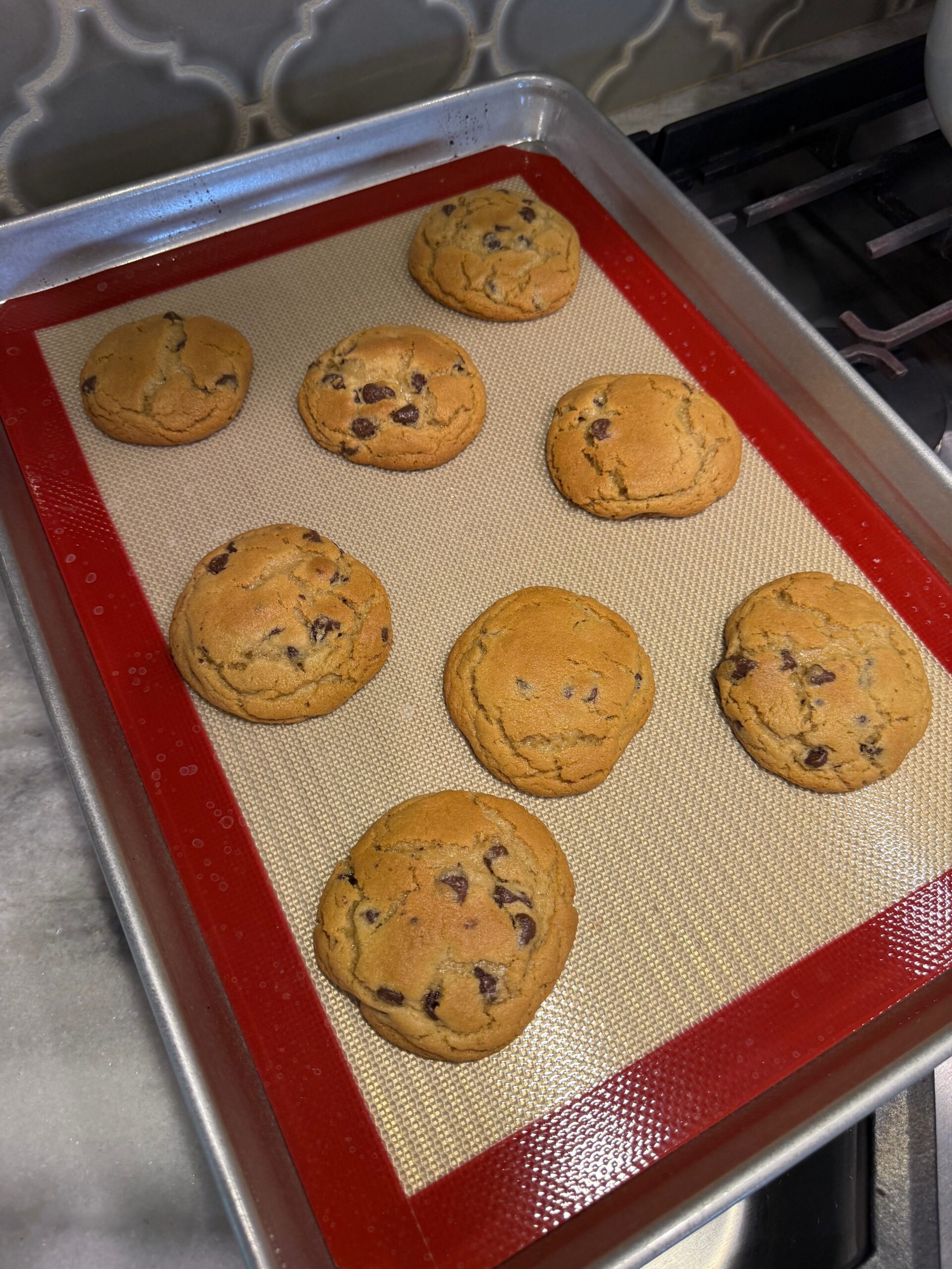 Nine chocolate chip cookies on a silicone baking mat-lined tray, fresh out of the oven, with a tiled backsplash and stove partially visible in the background.