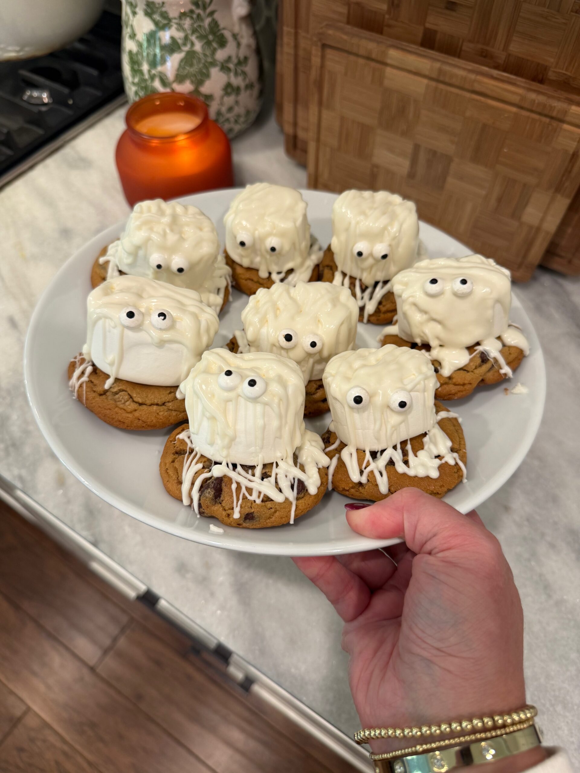 A hand holds a plate of cookies topped with marshmallows decorated as white, drippy ghosts with candy eyes. The plate is in a kitchen setting with a lit candle and wooden containers in the background.