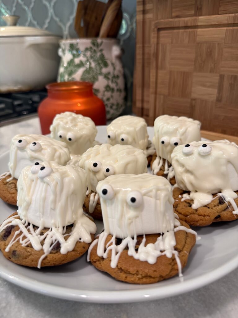 Chocolate chip cookies topped with marshmallows, white chocolate drizzle, and candy eyes to look like ghosts, arranged on a white plate in a kitchen setting with a red candle in the background.