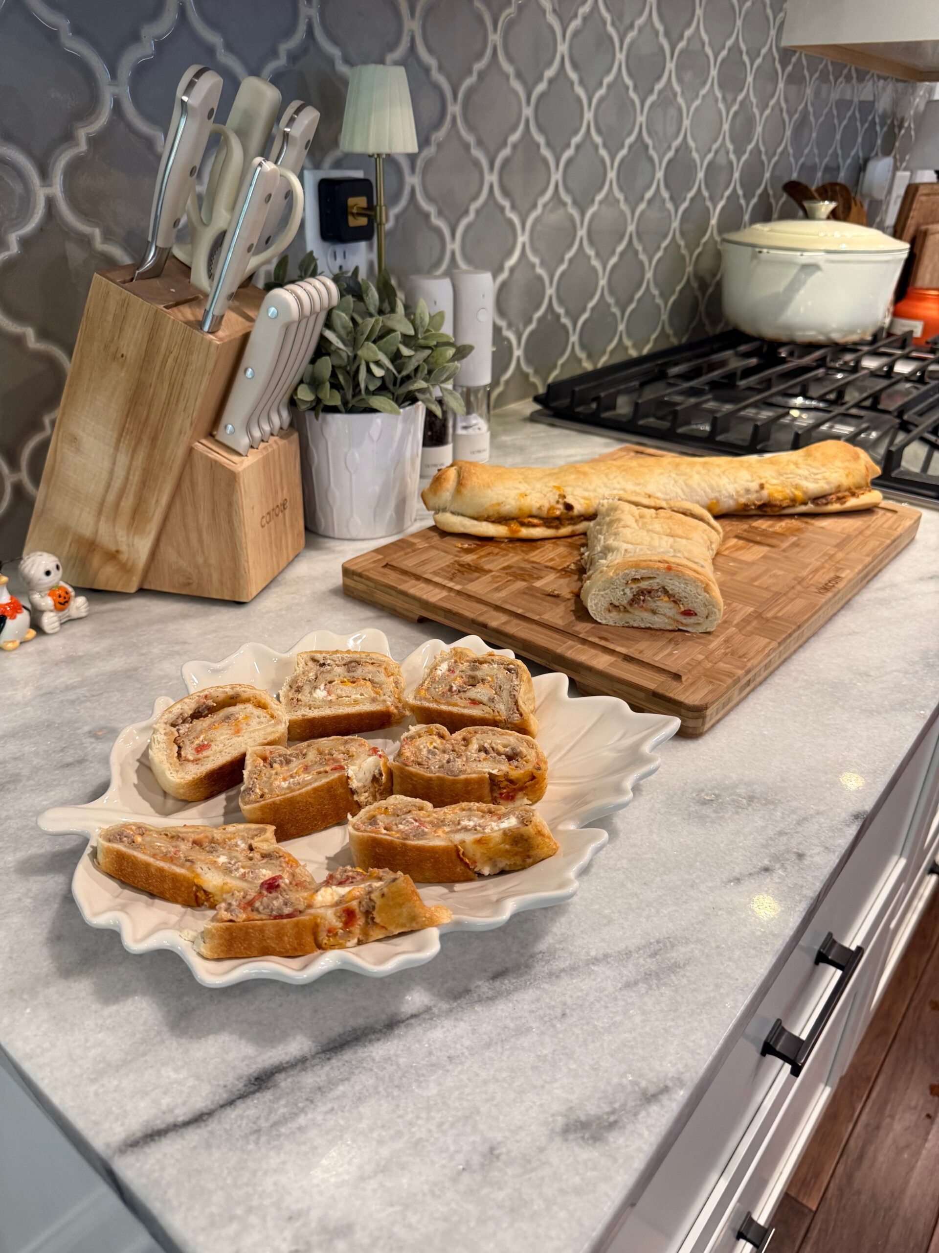 Sliced bread filled with meat and cheese is displayed on a decorative plate and wooden board on a kitchen counter. A knife block, plant, and stove are visible in the background.